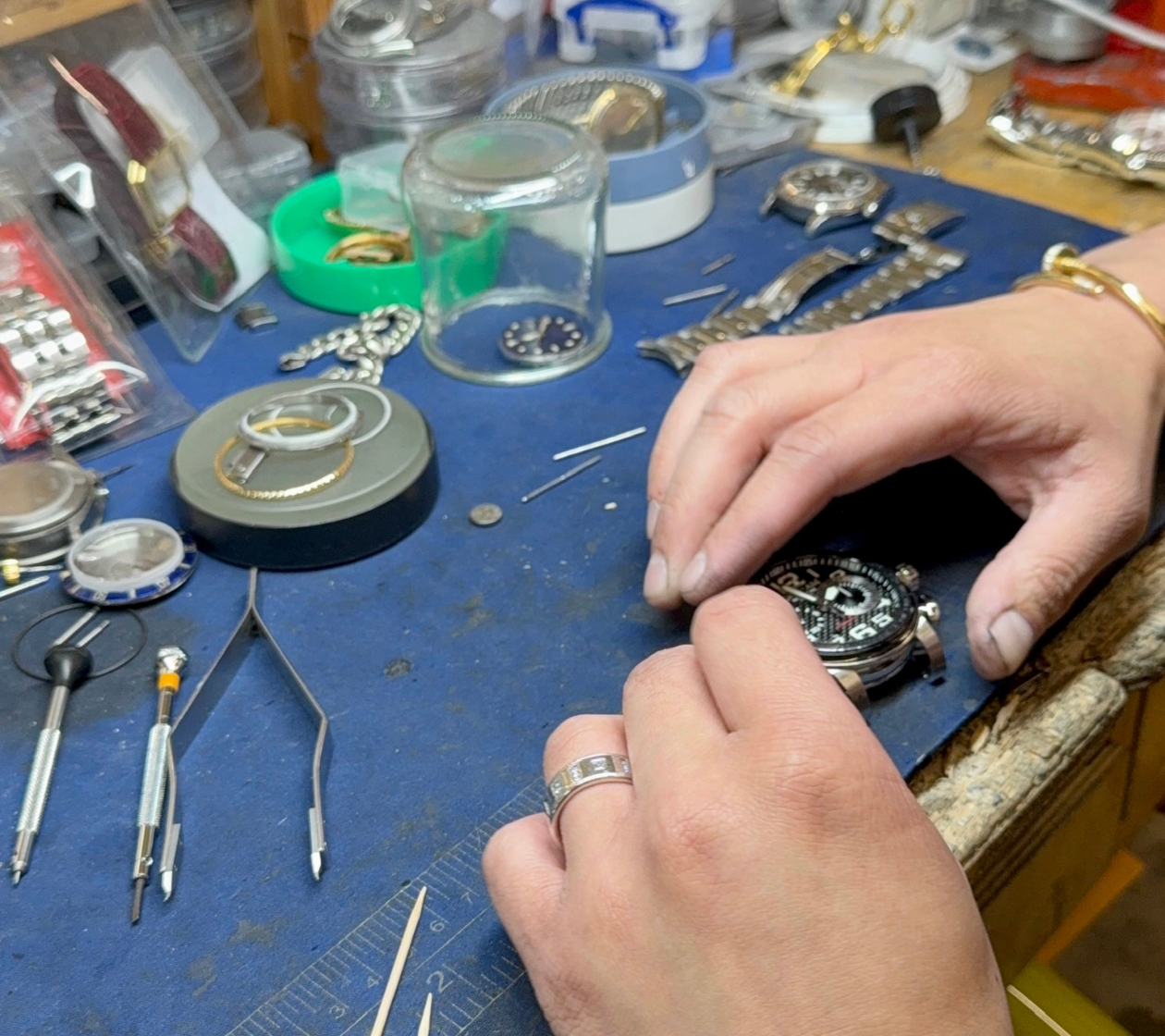 Watchmaker repairs a complex timepiece at a cluttered blue workbench with precision tools and parts nearby.