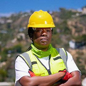 Construction worker wearing a yellow hard hat and high-visibility vest with arms crossed at a construction site.