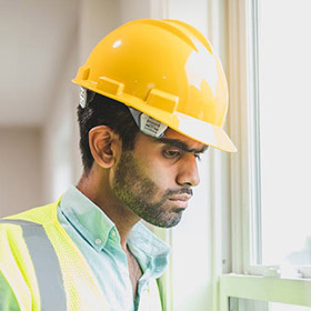 Construction worker wearing a yellow hard hat and reflective vest, looking out a window.