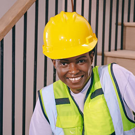 Smiling construction worker in a yellow hard hat and neon safety vest indoors near a staircase.
